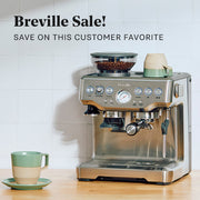 Breville Barista Express in Stainless Steel on a wooden counter top in front of a white tiled wall next to a sage cup. Promotional text overlayed that reads "Breville Sale! Save on this customer favorite".