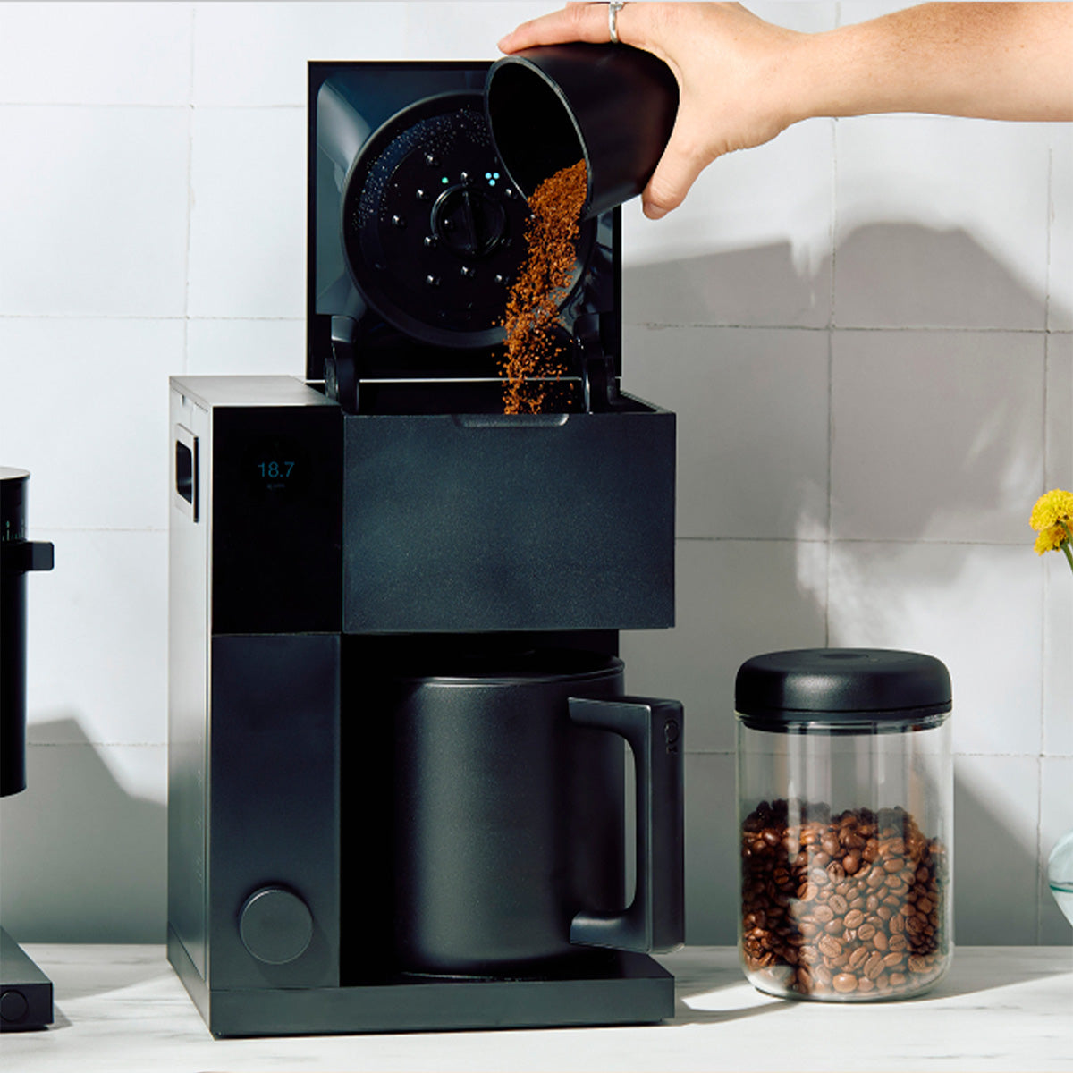 Person pouring coffee grounds into a coffee maker with a container of coffee beans on a kitchen counter.