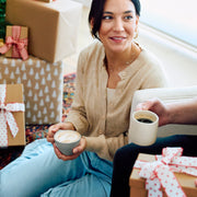 Woman sitting on a couch holding coffee cups with Christmas presents in the background