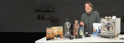 Marc from Whole Latte Love standing behind a display of coffee-making equipment, like grinders, coffee, coffee accessories and a Synchronika II with a dark background