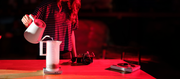 Close-up of a person in a striped shirt pours hot water from a white pitcher into a white carafe set on a scale on a wooden table, illuminated by dramatic red lighting in a dark room.