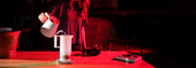 A person in a striped shirt pours hot water from a white pitcher into a white carafe set on a scale on a wooden table, illuminated by dramatic red lighting in a dark room.