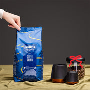 Person holding bag of Lavazza Super Crema Coffee next to coffee cups and canisters in front of a black wall.