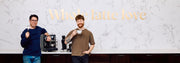 Two men holding coffee cups in front of a coffee machine with 'Whole latte love' text on a white marble wall.