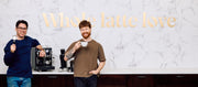Close-up of two men holding coffee cups and portafilter in front of a coffee machine with 'Whole latte love' text on a white marble wall.