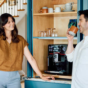 Two people in a kitchen with a Gaggia Accademia espresso machine and shelves holding cups and glasses.