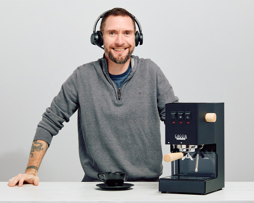 Man with headphones next to a Gaggia Classic Pro Espresso Machine and cup on a light gray background