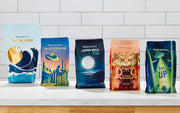 Line-up of Whole Latte Love roasted coffees on a white counter in front of a white tiled backsplash.
