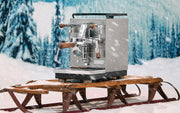 Profitec JUMP Espresso machine on a wooden sled in a snowy landscape with trees and snow falling.