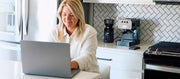 Close-up of a woman using a laptop in a kitchen with Gaggia Espresso Evolution on the counter behind her. 
