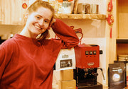 A smiling woman in a red sweatshirt stands next to a red Gaggia Classic Pro E24 espresso machine and bags of coffee..