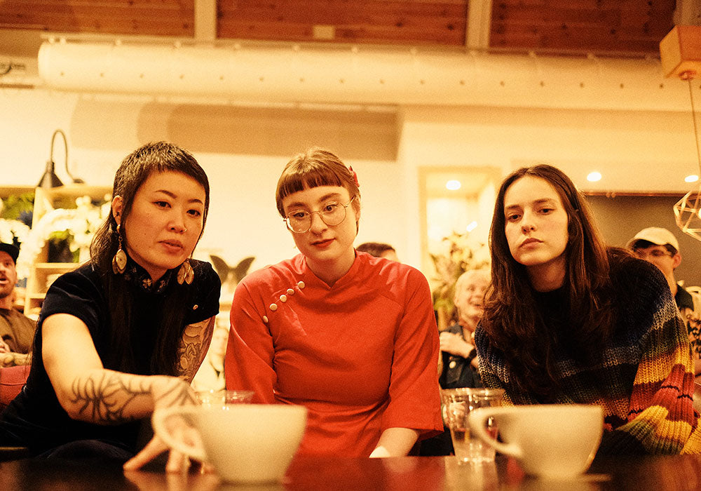 Three women latte art judges seated at a table in a warm, dimly lit indoor setting, with white cups in the foreground.