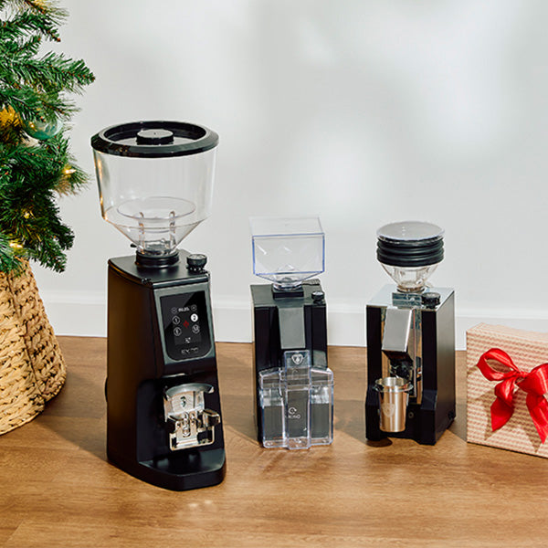 Three coffee grinders on a wooden surface with a Christmas tree and gift box in the background.